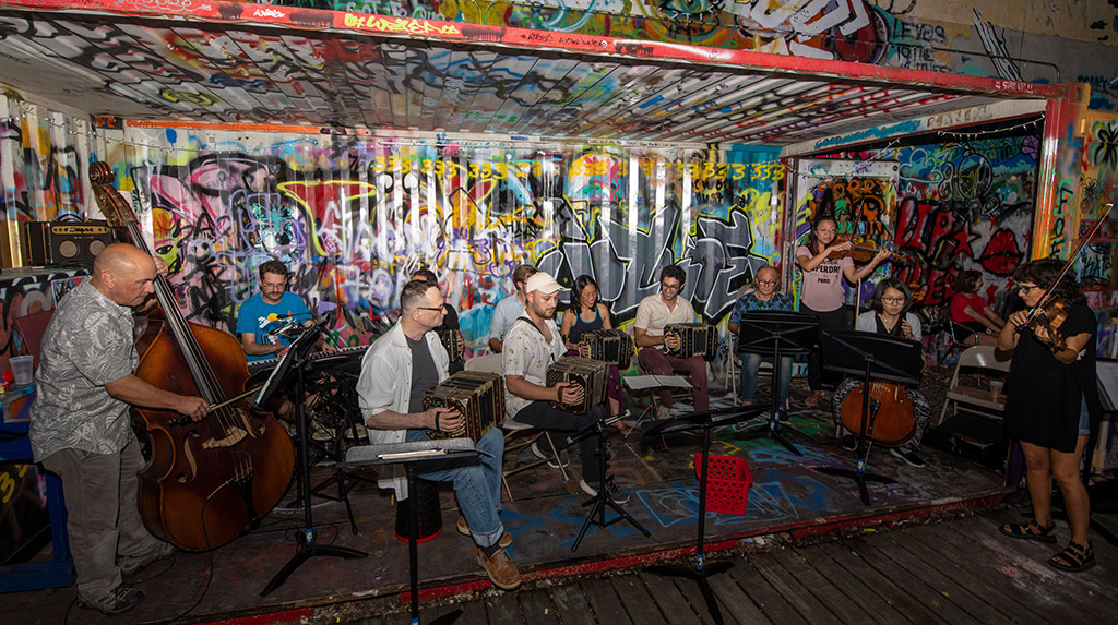This is a photo of a group of musicians playing in front of a colorful graffiti wall. The musicians are playing various instruments, including an accordion, a guitar, and a drum set. The graffiti wall is covered in vibrant colors and abstract designs. The musicians are sitting on a green carpeted floor. The image is slightly blurred, likely due to the movement of the musicians.