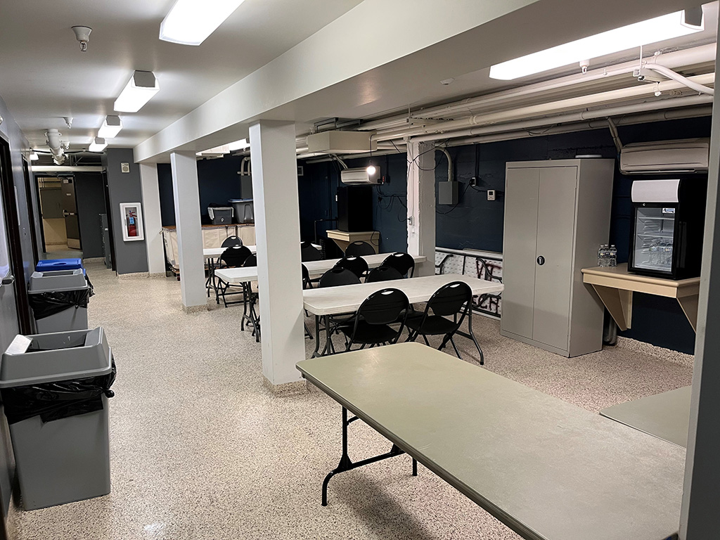A room with white ceiling and white pillars with blue walls. There are several rectangular folding tables set up with folding chairs. There is a mini-fridge, a filing cabinet and several garbage cans. There is a lot of fluorescent lighting.