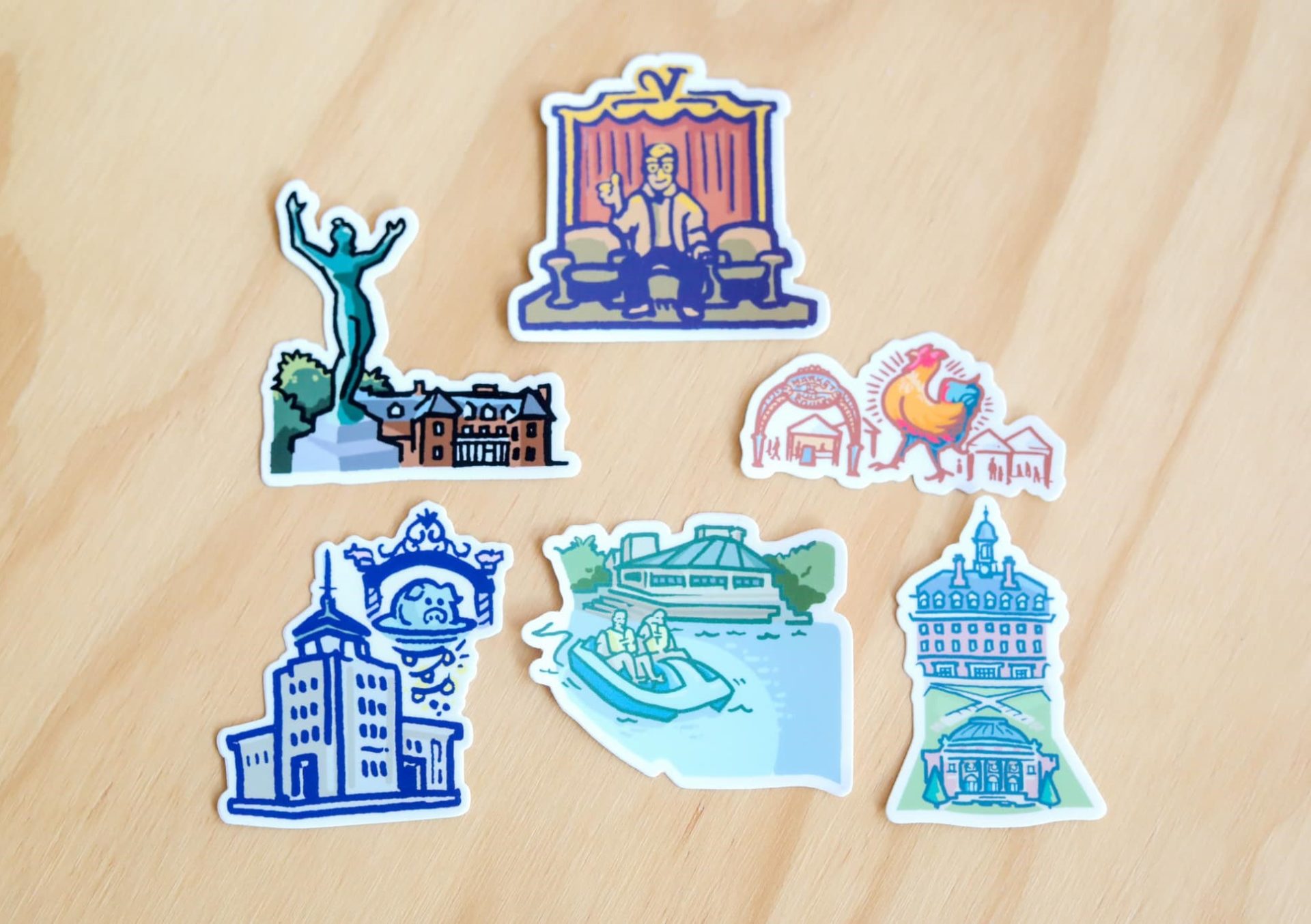 A collection of five stickers with artwork depicting Champaign-Urbana related things. The are arranged in a cluster on a light wood surface.