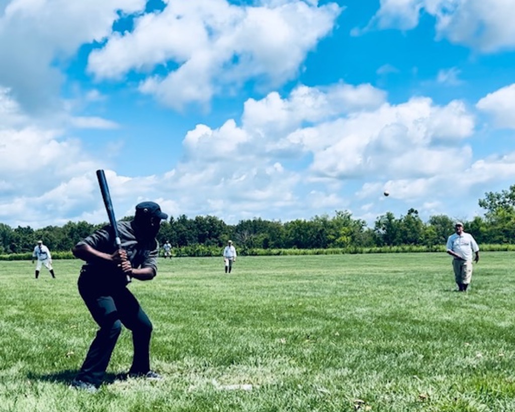 A black man in dark pants, and a shirt stands on a grassy field with a blue sky with fluffy white clouds. In the foreground of the photo hi is in a batting stance, getting ready to swing a bat. 