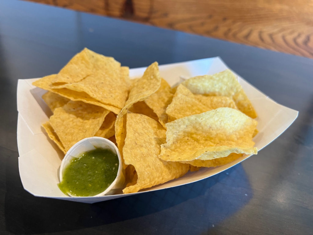 A white paper basket of tortilla chips and green salsa is on a black table. Photo by Alyssa Buckley