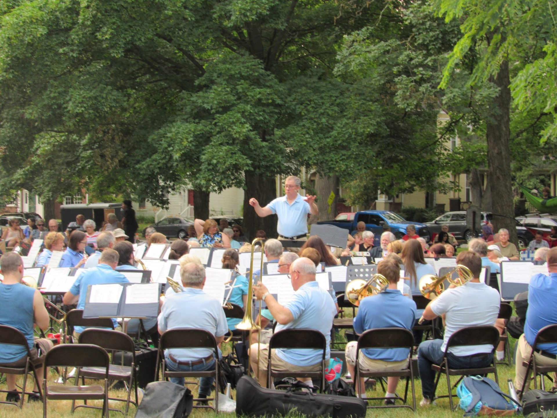 A concert band arranged in folding chairs in a park. The musicians are all wearing light blue shirts. A white man, also in a light blue shirt, is standing in front conducting.