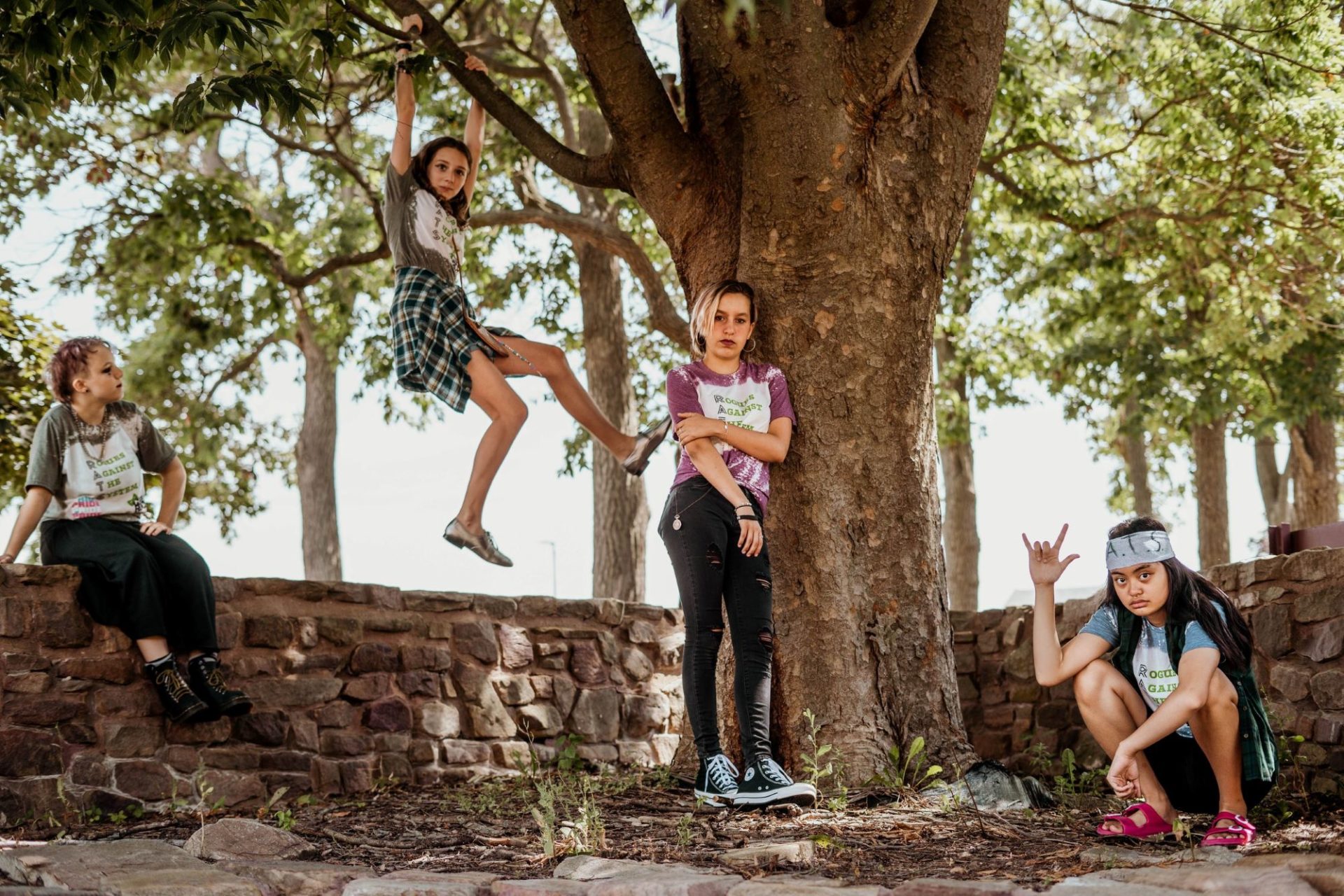 Four young teens are posing near a large tree, surrounded by a low stone wall. One is sitting on the wall, one is hanging from by her hands from a branch, one is leaning against the tree, one is crouched on the ground.