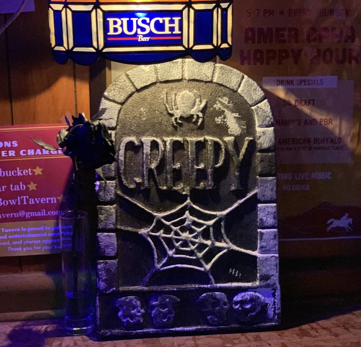 A fake tombstone that says "creepy" and has a spider web and spider on it sits on a counter at a bar. A Busch beer lamp hangs over it.