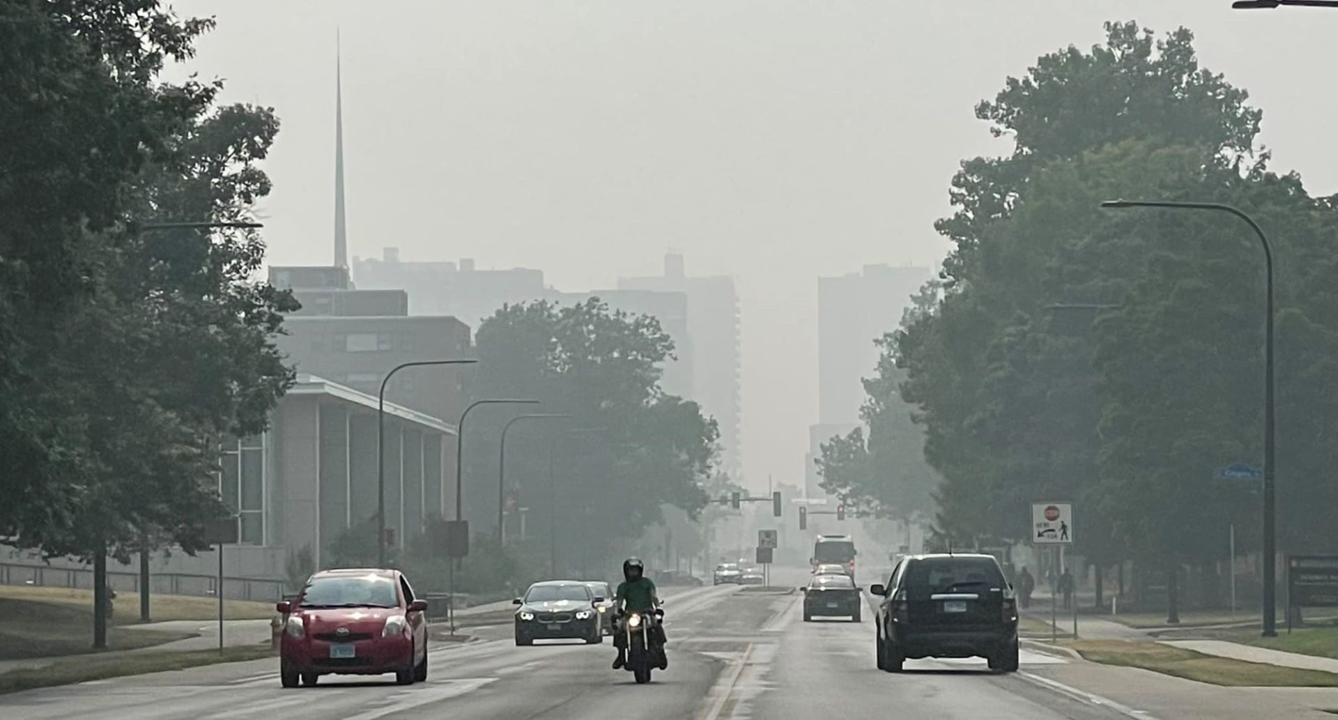 View down a campus street with buildings and trees lining the road. There are cars traveling in each direction. The air is gray and very hazy.