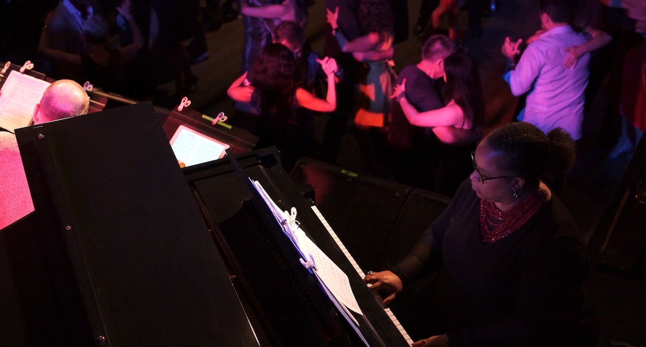 A photograph from above of a black female pianist with tango dancers dancing behind her. The lighting is dark with some pink tones.