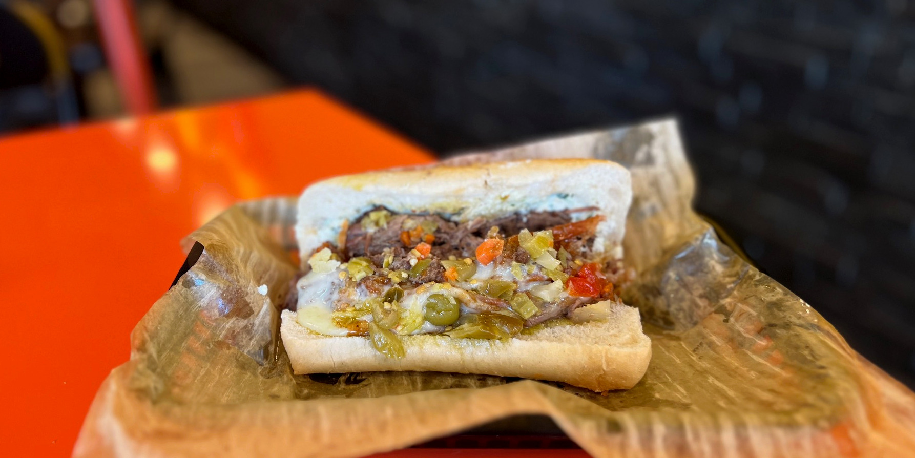 A cropped image of Dee-Dee Beef's Italian beef sandwich on brown parchment paper in a black plastic basket on an orange table. Photo by Alyssa Buckley.