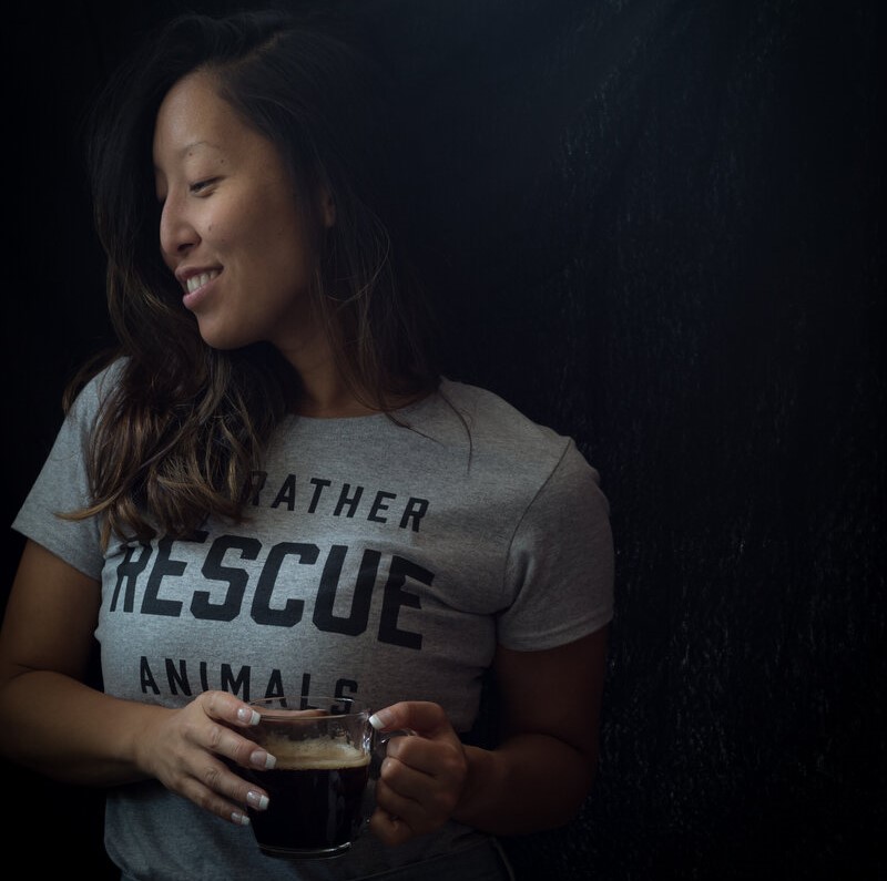 An Asian woman with long brown hair, wearing a gray t shirt, is standing and holding a clear mug of coffee. She is looking off to the side and smiling.