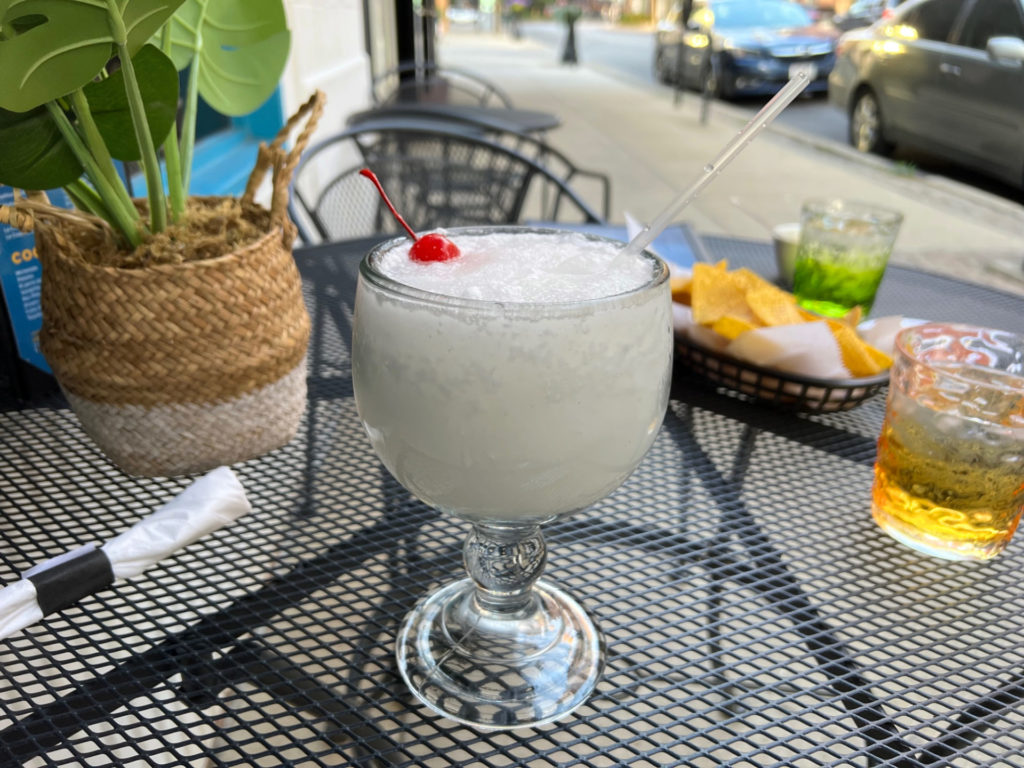 A piña colada margarita drink with a cherry on top sits on a black patio table outside La Bahia Grill for summer eats in Downtown Champaign. Photo by Alyssa Buckley.