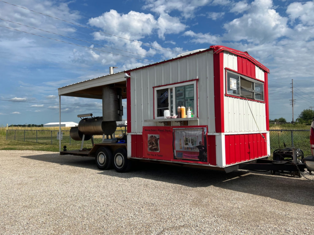Mama Duke's red and white trailer is parked at Riggs in Urbana. The trailer has a barbecue smoker on the back "patio" of the trailer. The sky is blue with fluffy white clouds. Photo by Alyssa Buckley.