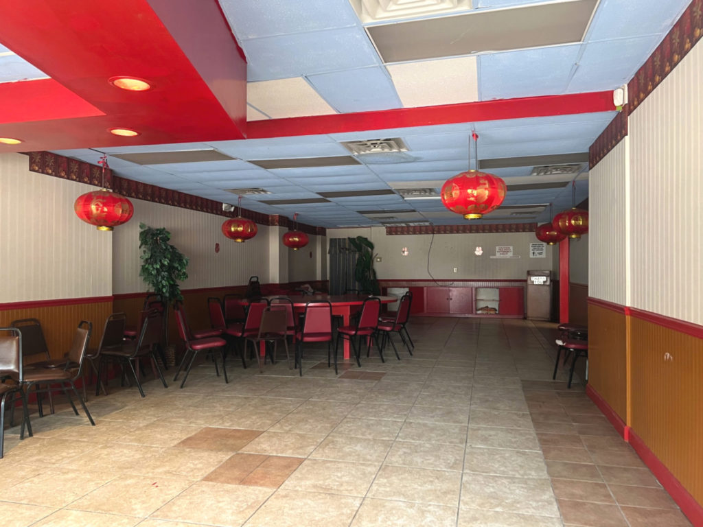 The interior of Mandarin Wok, now closed, as seen through the window. There is one large red table and a few haphazard chairs with an empty dining room. Photo by Alyssa Buckley.