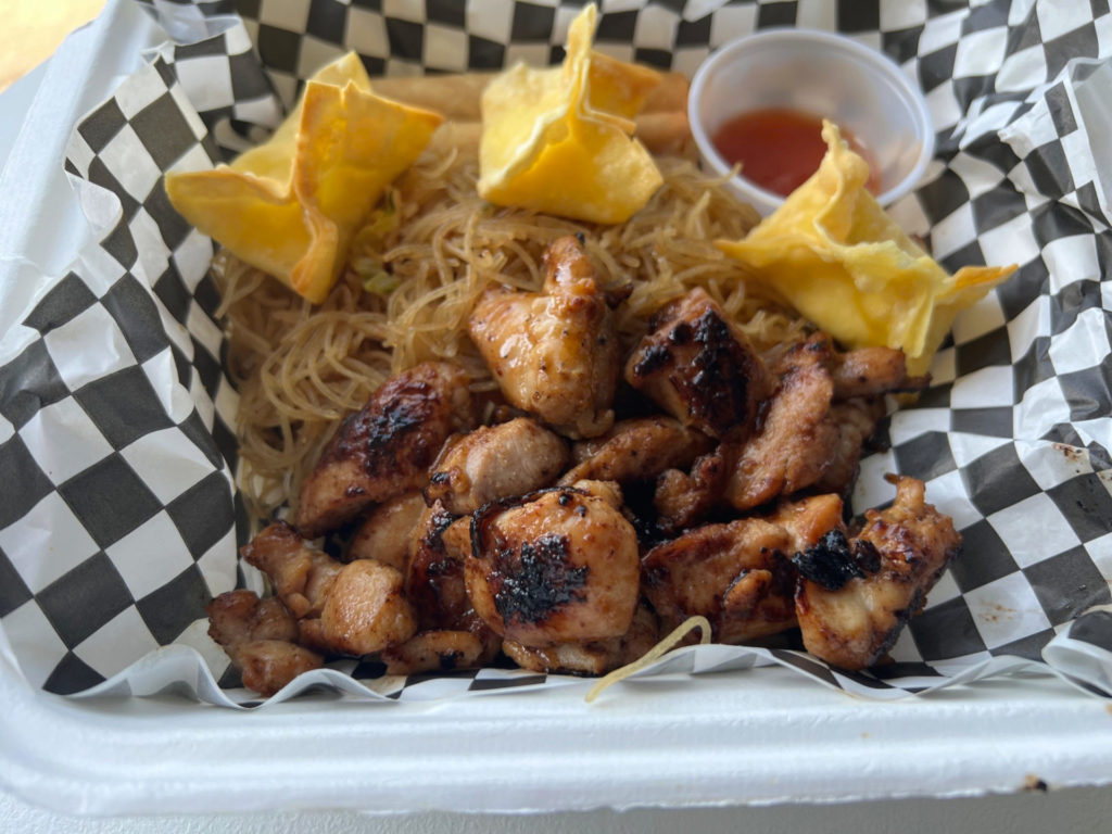 A takeout container from Maria's Kitchen food truck is lined with a black-and-white parchment paper with grilled chicken, pancit noodles, and three small rangoons with a cup of sauce. Photo by Alyssa Buckley.