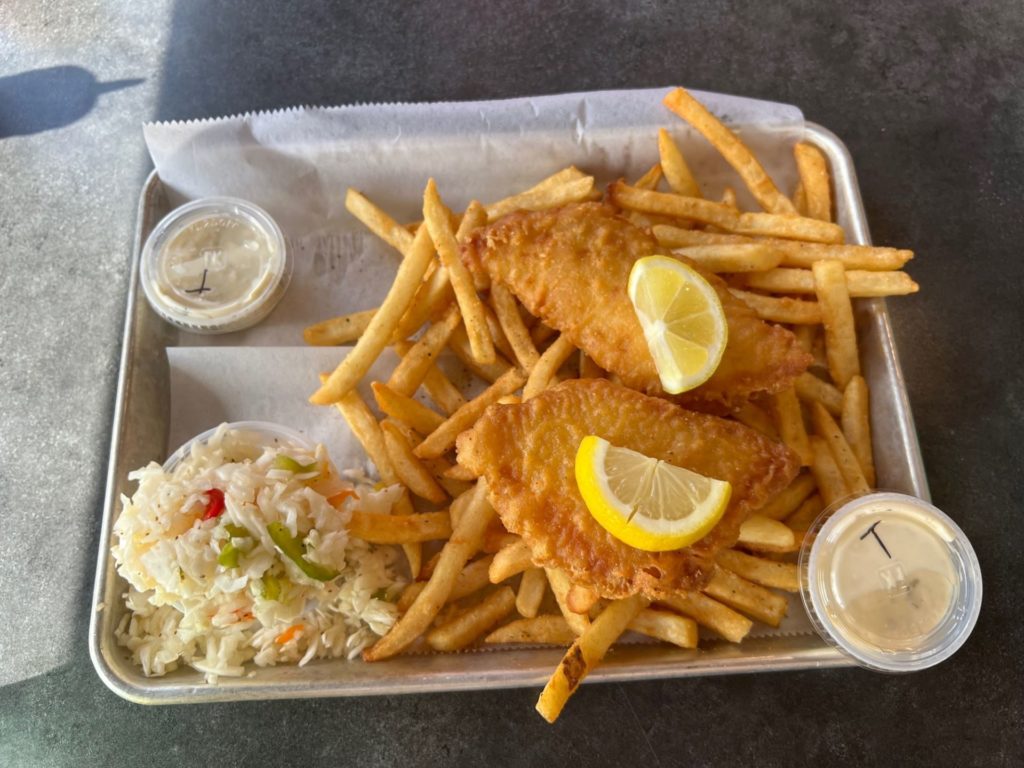 A metal tray, lined with wax paper. It's piled with french fries and two breaded fish fillets. There are two plastic cups of creamy sauce, and a larger plastic cup with coleslaw.