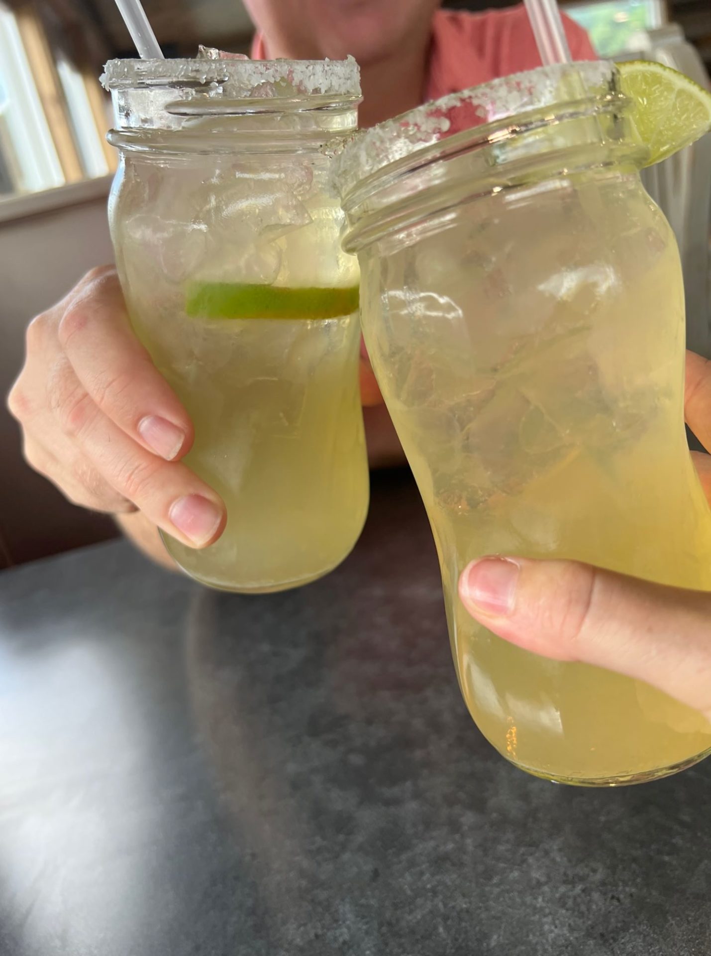 A close up of hands, each holding a glass with a yellowish liquid. The glass rims are lined with salt, and have a lime on the edge.