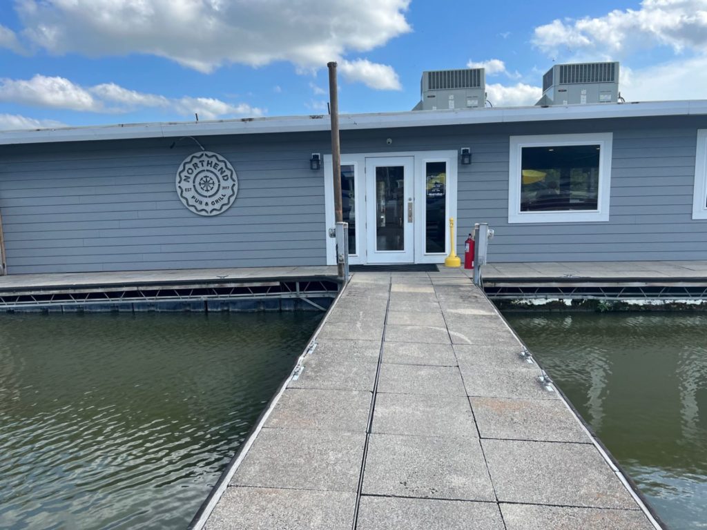 A dock with cement tiles leads up to a small building with bluish gray siding. It has a circular metal sign that says North End Pub & Grill.