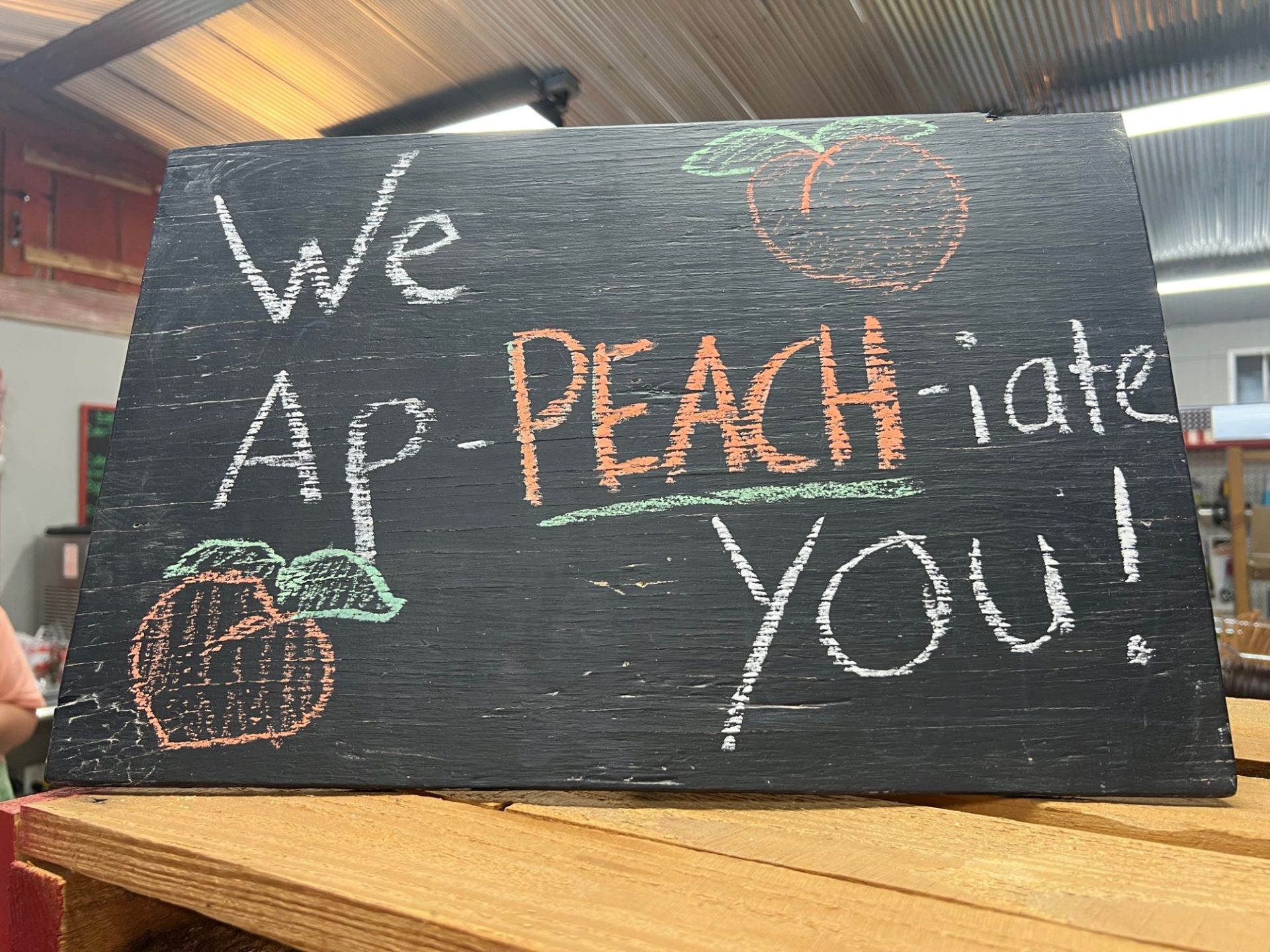 A chalkboard sign, sitting on a wooden shelf. It says We Ap-Peach-iate You in chalk.