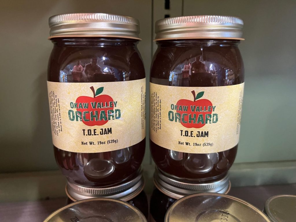 Two large glass jars with a brown-ish jam inside of them. They have beige labels with a red apple, and Okaw Valley Orchard in green block letters.