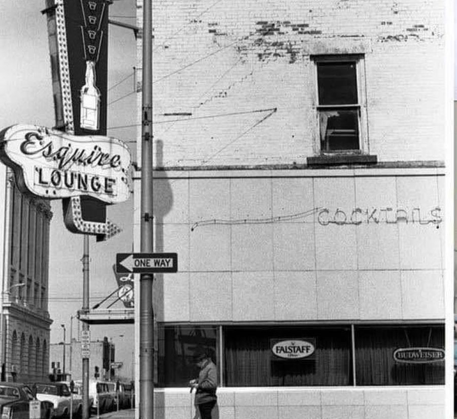 Black and white photo of a building on a street corner with a neon sign hanging on the corner of the building, that says Esquire Lounge.