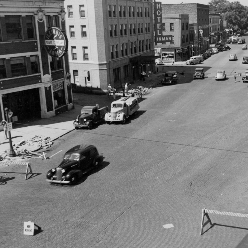 A black and white photo of a city block, with old-fashioned cards driving on the road. There are brick building lining the road. One building has a sign that says Hotel Inman jutting out from the corner.