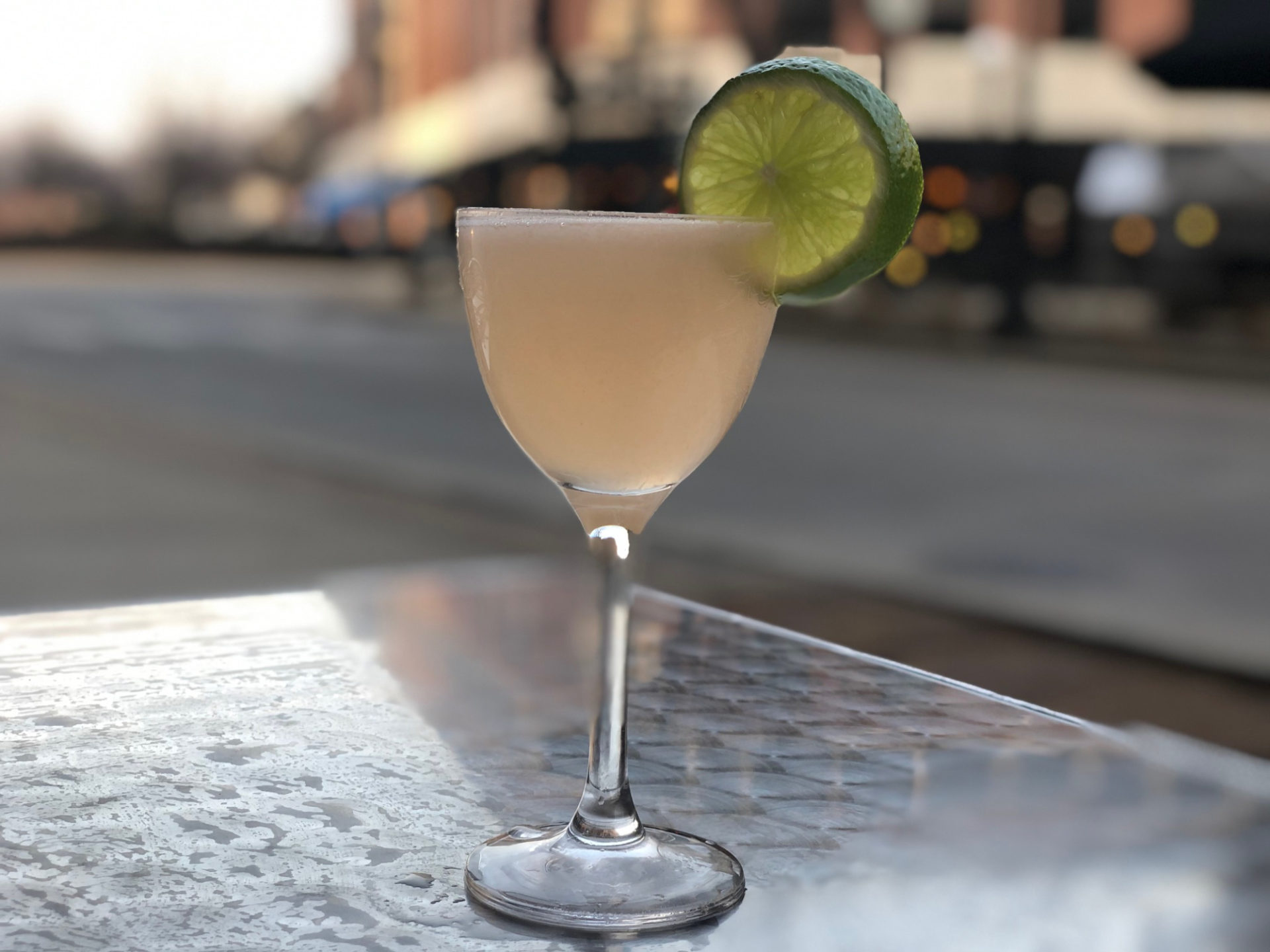 A light-colored drink in a stemmed glass, with a lime garnish, is sitting on a metal table. There is a street and buildings in the background.