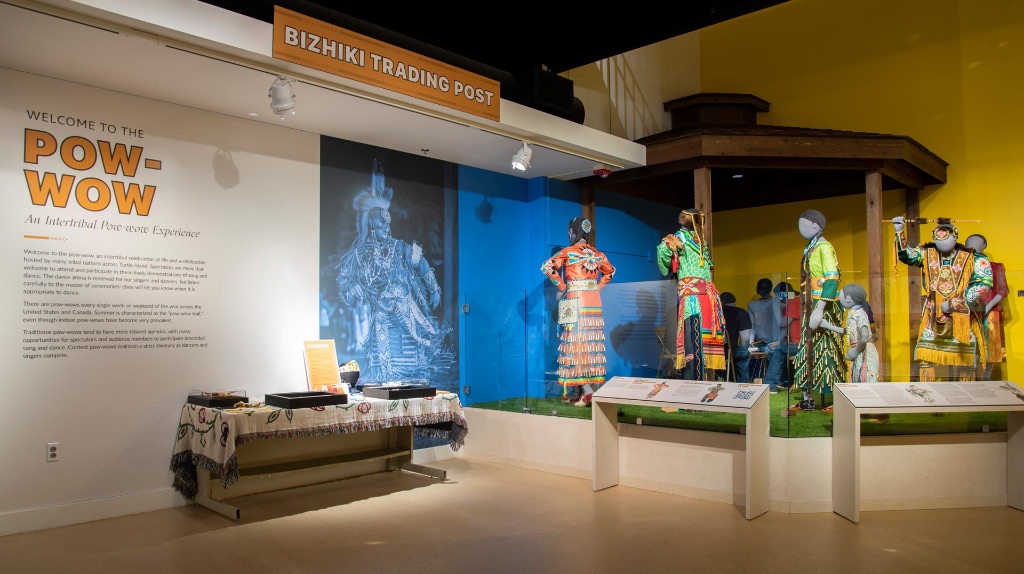 a museum exhibit featuring different pow wow costumes and headdresses on mannequins. Behind them there is a wall featuring large pictures and text describing the exhibit. 