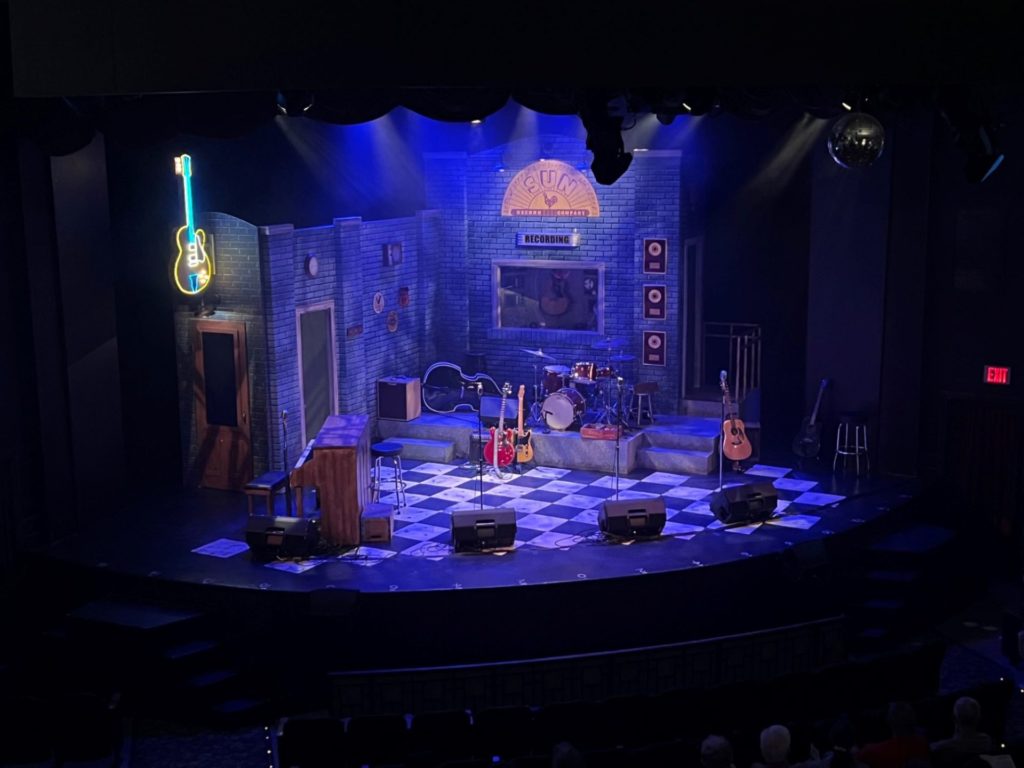 A stage set up to look like the inside of a recording studio. There are microphones, guitars, a piano, drumset, and large upright bass on the stage, that has a black and white checkered floor.