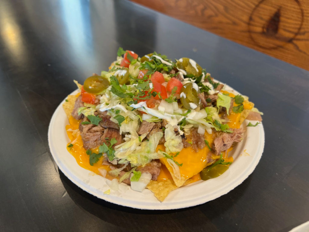 A white paper plate of La Paloma nachos piled high ith carnitas, orange nacho cheese, lettuce, onions, tomato, sour cream, tomatoes, and jalapenos. Photo by Alyssa Buckley.