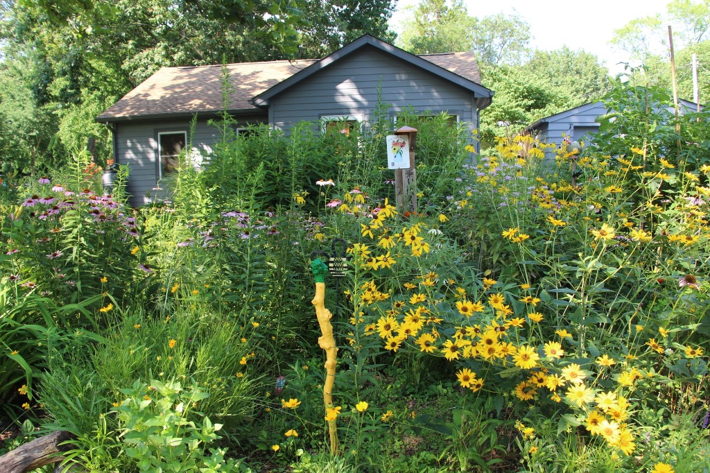 A garden full of flowers and other tall green plants takes over most of the image. In the foreground is a stick painted yellow and green. In the background is a teal colored house with only the very top and roof line visible behind the plants.