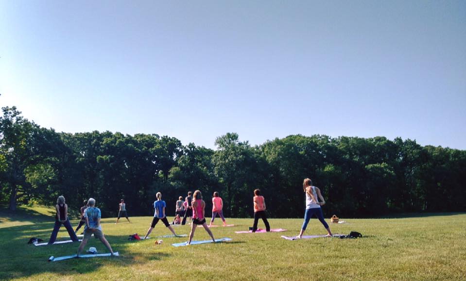 A grassy meadow on a sunny day features a group of woman in yoga pants and bright tops standing on Yoga mats.