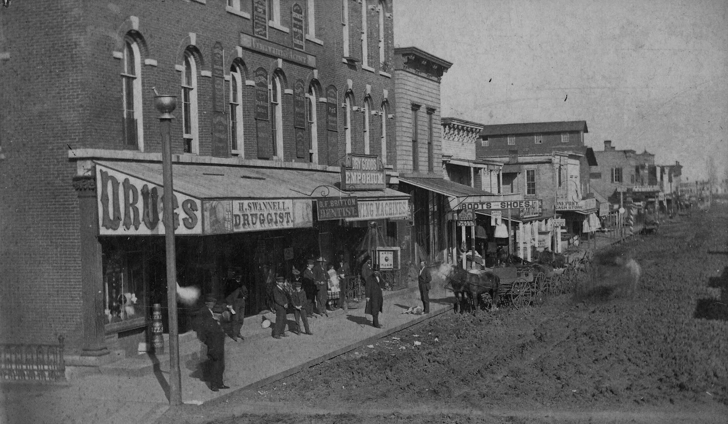 A black-and-white photo of 1 East Main Street in Champaign, Illinois. There is a dirt road with horses, and men in top hats on the sideway. Photo credit to Champaign County Historical Archives at The Urbana Free Library.