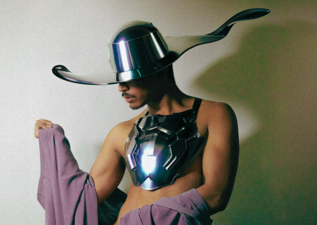 Marcus Flinn is shirtless with a chrome breastplate with mauve fabric draped over his arms. He is looking down to the left and has a large brimmed chrome hat. 