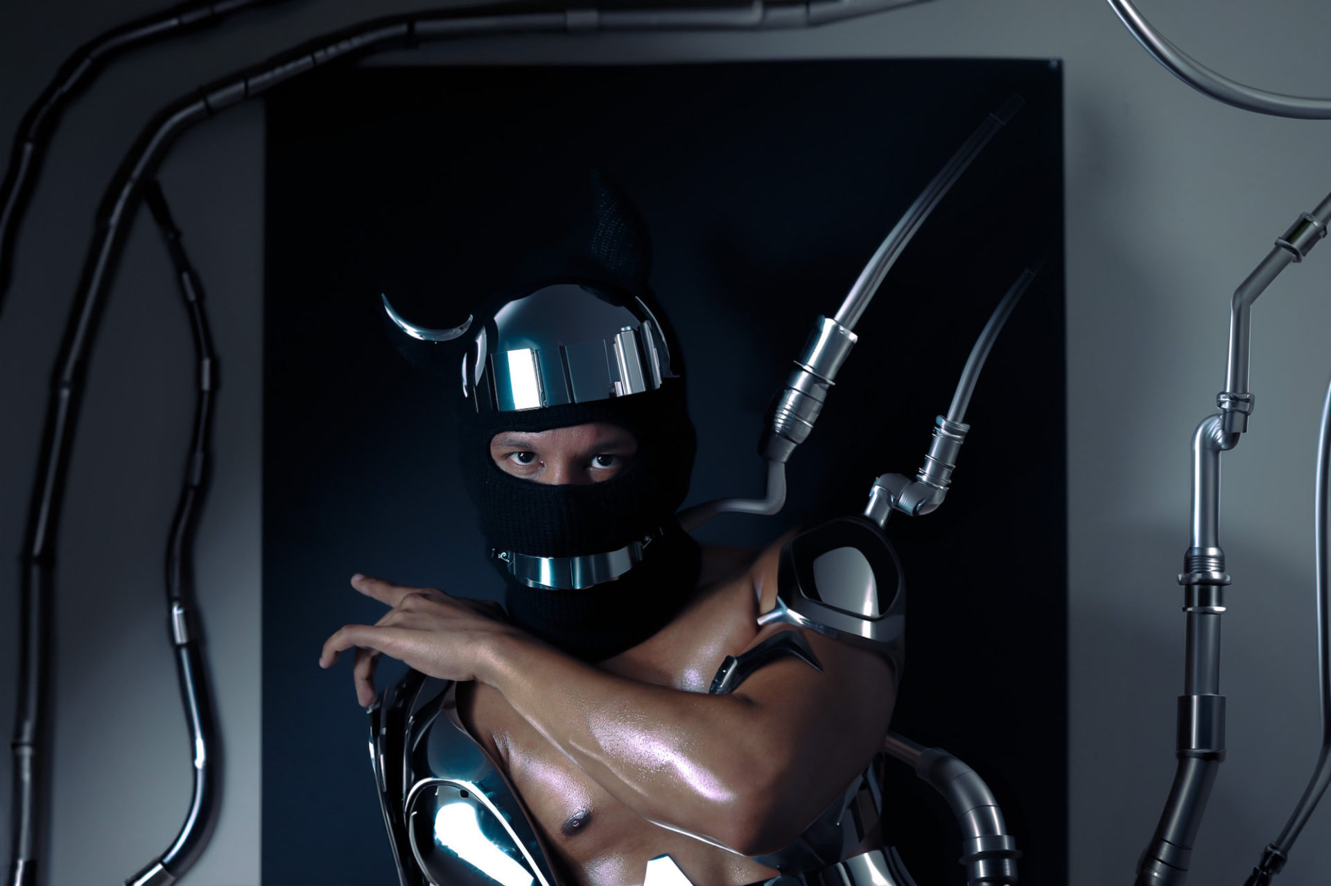 Portrait by Marcus Flinn. He is shirtless, with a chrome helmet and wearing large chrome futuristic pieces on his arms. The photo is mostly grey and black. He is standing in from of a black backdrop, with grey and tubing framing him
