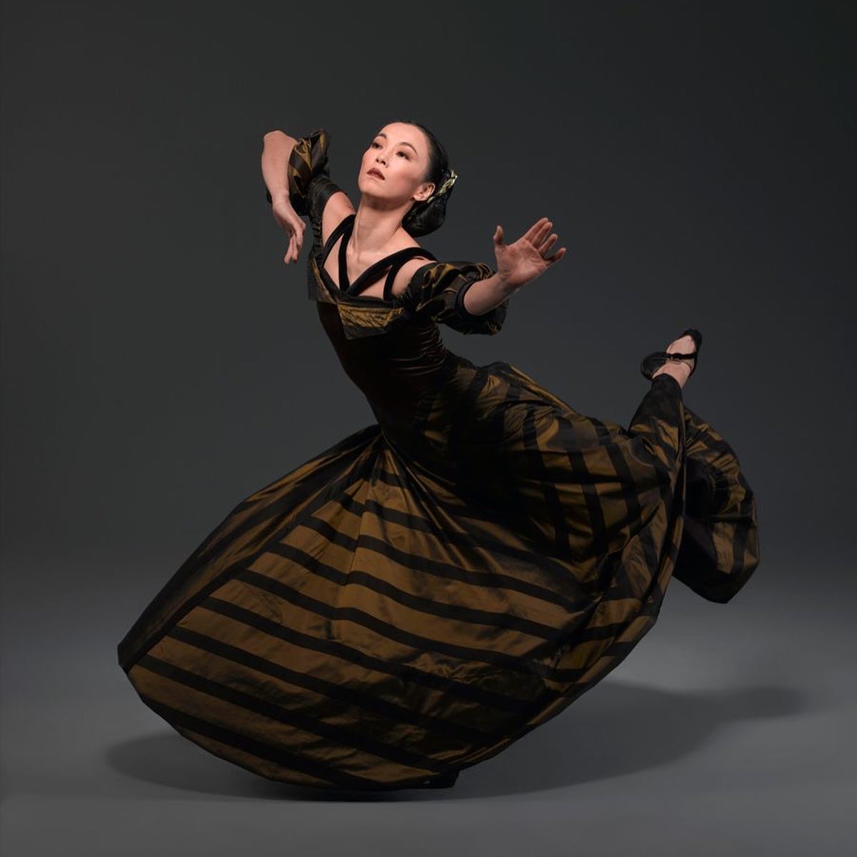 Miki Orihara, a Japenese woman, has her black hair pulled into a low bun. She is standing on one foot with one leg bent and extended behind her. She is holding her arms in a dance hold. She is wearing a floor length flamenco-style black and beige stripped tulle gown.