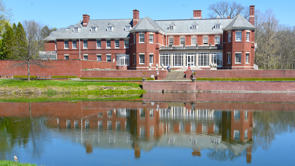 The Allerton Park mansion viewed from the back. It is a huge red brick mansion with white windows and a gray roof. The are retaining walls and steps that lead to a pond, in which you can see the mansion reflected. It is a sunny day with a light blue sky.