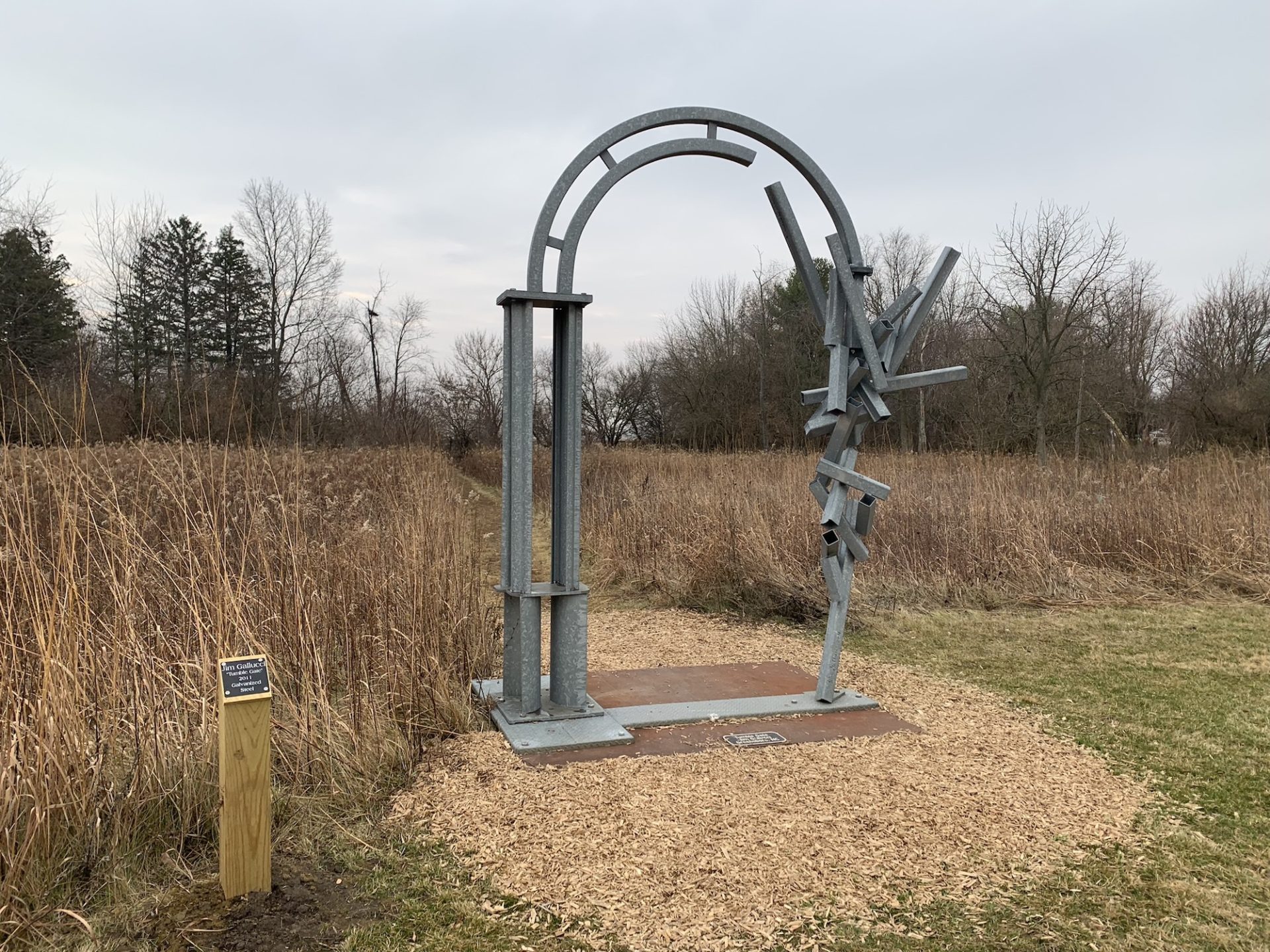 A large metal arched sculpture stands in a park. The left side of the arch has straight columns, while the right side has beams assembled going in every direction creating a 3-D effect.