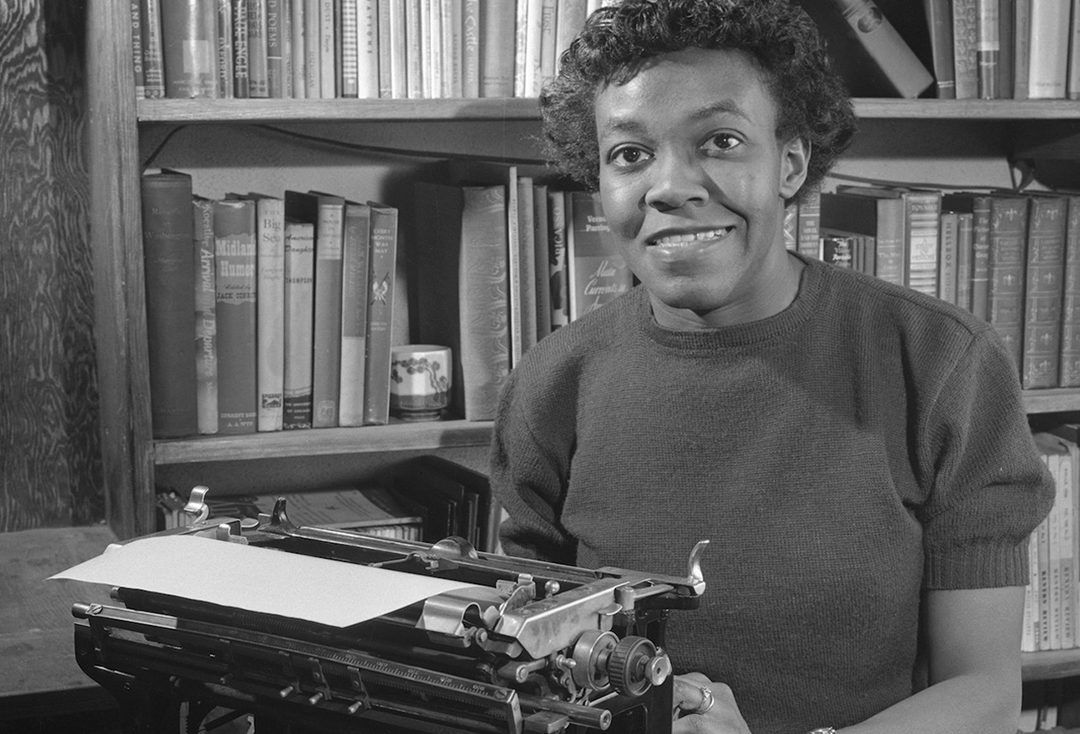 black and white photograph of Gwendolyn Brooks, a Black woman with short black hair. She sits in fron t of a wall of books, wearing a short sleeved sweater and sitting in front of a typewriter