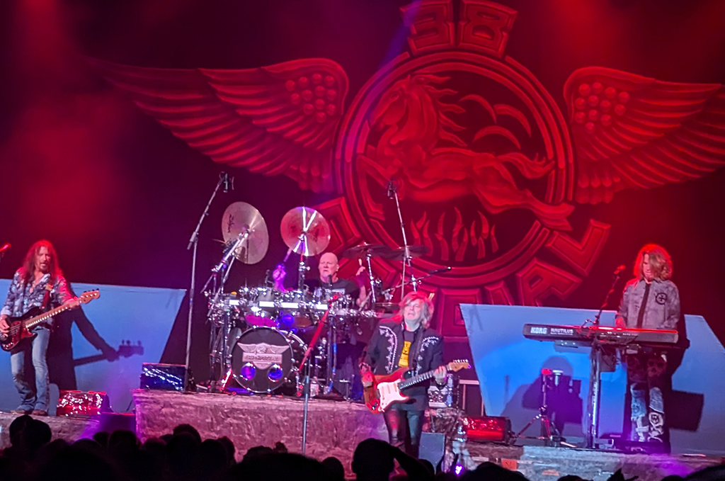 This is a photo of a rock band performing on stage. The stage is decorated with a large red and gold emblem of a winged eagle with a drum set in front of it. There are four band members visible on stage, each playing a different instrument. The band members are dressed in casual clothing and have long hair. The stage is lit with red and purple lights, creating a dramatic and energetic atmosphere. The audience is not visible in the photo.
