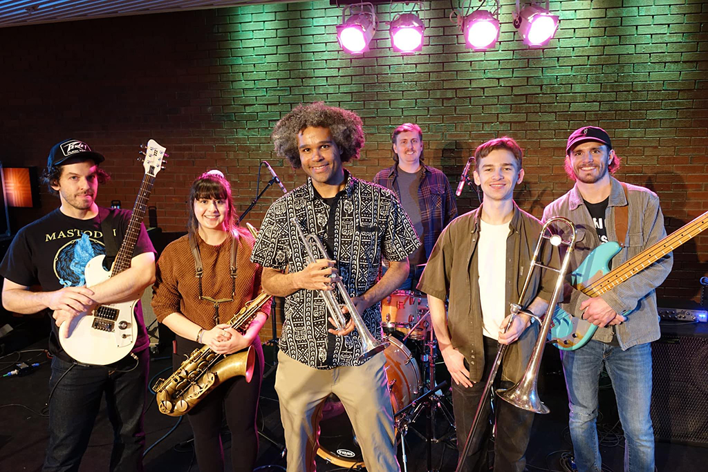 This is a photo of a band on stage. The band consists of six members, each holding a different instrument. The instruments include a guitar, a saxophone, a trumpet, a trombone, a bass guitar, and a drum set. The band members are standing in front of a brick wall with green stage lights shining down on them. The band members are wearing casual clothing, with one member wearing a Mastodon t-shirt.