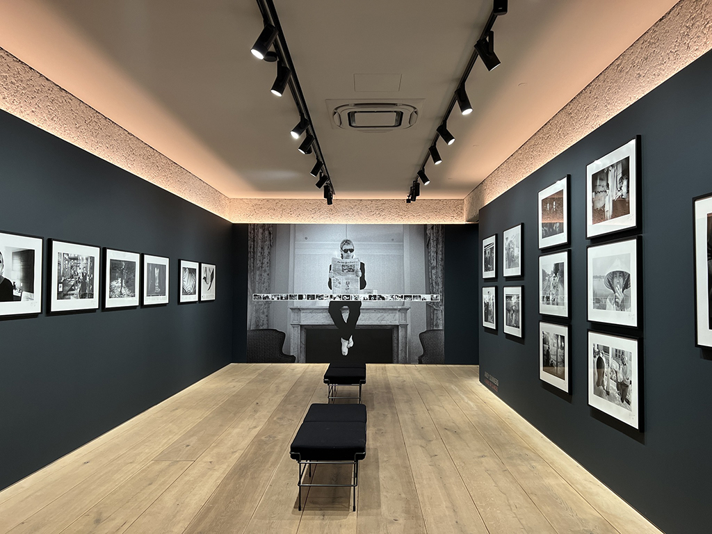 This is an image of an art gallery with black and white photographs on the walls. The gallery has a wooden floor and a black bench in the center. The walls are painted dark gray and the ceiling is white with track lighting. The photographs are framed in black and are of various sizes. The largest photograph is on the back wall and is of a person with their arms outstretched holding a newspapers sitting on a fireplace mantle. The other photographs are of people, buildings, and street scenes.