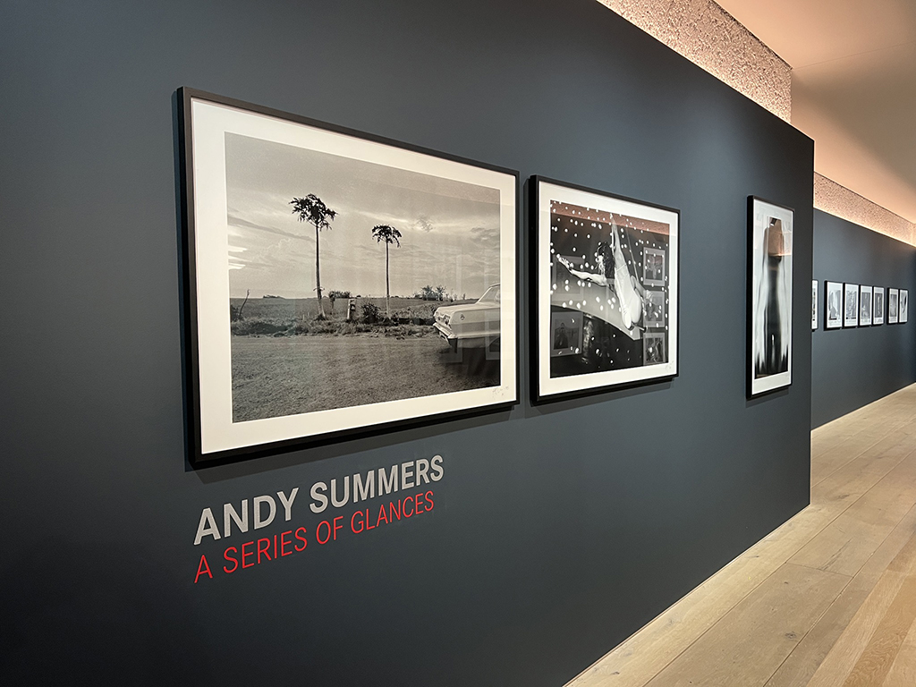 This is a photo of an art gallery wall with three framed black and white photographs. The left photograph is of a beach with palm trees and a boat, the middle photograph is of a car parked on a street, and the right photograph is of a city street at night with lights. The photographs are hung on a gray wall with red text that reads “ANDY SUMMERS A SERIES OF GLANCES”.