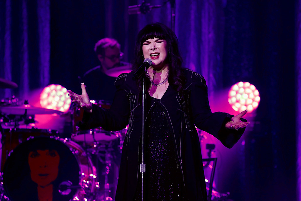 This is a photo realistic image of a singer on stage. The singer is wearing a black jacket with a purple shirt underneath. They are standing in front of a drum set and a microphone. The background consists of a stage with purple lighting and a large screen with a face on it.