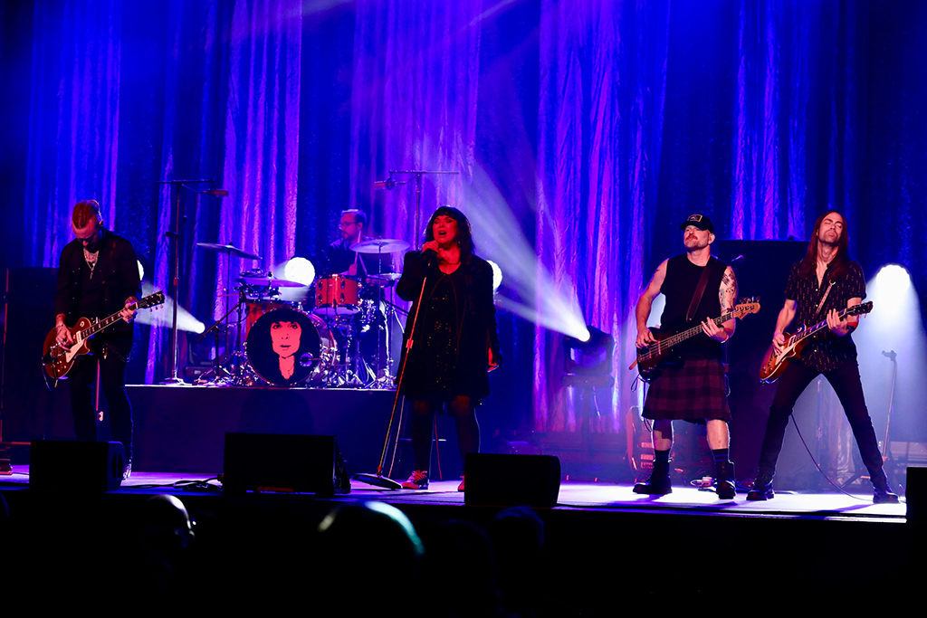 This is a photo of a rock band performing on a stage. The stage is lit with blue and purple lights. The band members are playing guitar, bass, drums, and singing. hey are wearing black clothing and one member is wearing a kilt. The background consists of a curtain and stage lights.