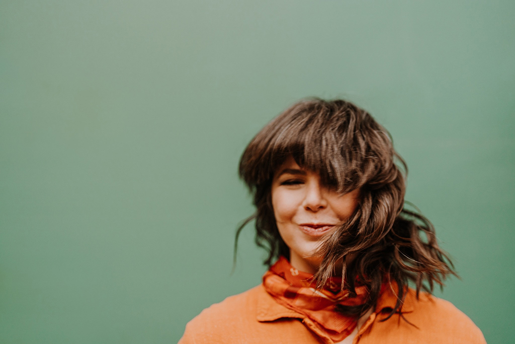 This is a photo of a person wearing an orange shirt and a scarf around their neck. The background is a solid green color. The person’s hair is brown and shoulder length. The image is cropped to only show the person’s upper body.