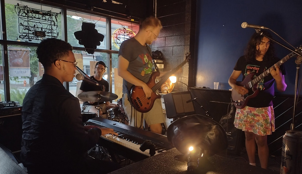 This is a photo of a band performing on a small stage in a bar. The band consists of a drummer, a guitarist, a bassist, and a keyboard player. The drummer is sitting behind a drum kit, the guitarist is playing a red electric guitar, the bassist is playing a white electric bass, and the keyboard player is playing a black keyboard. The stage is lit with blue and orange lights. The background consists of a window with a neon sign and a brick wall. The foreground consists of a table with a black helmet on it.