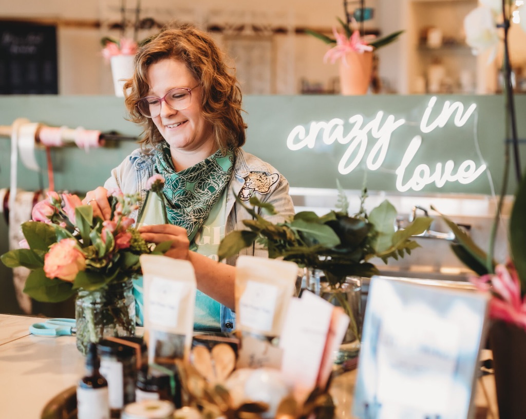 a white woman with shoulder length hair arranges flowers in front of a counter with cards and other gifts. She is wearing a green scarf there is a neon sign behind her that says crazy in love