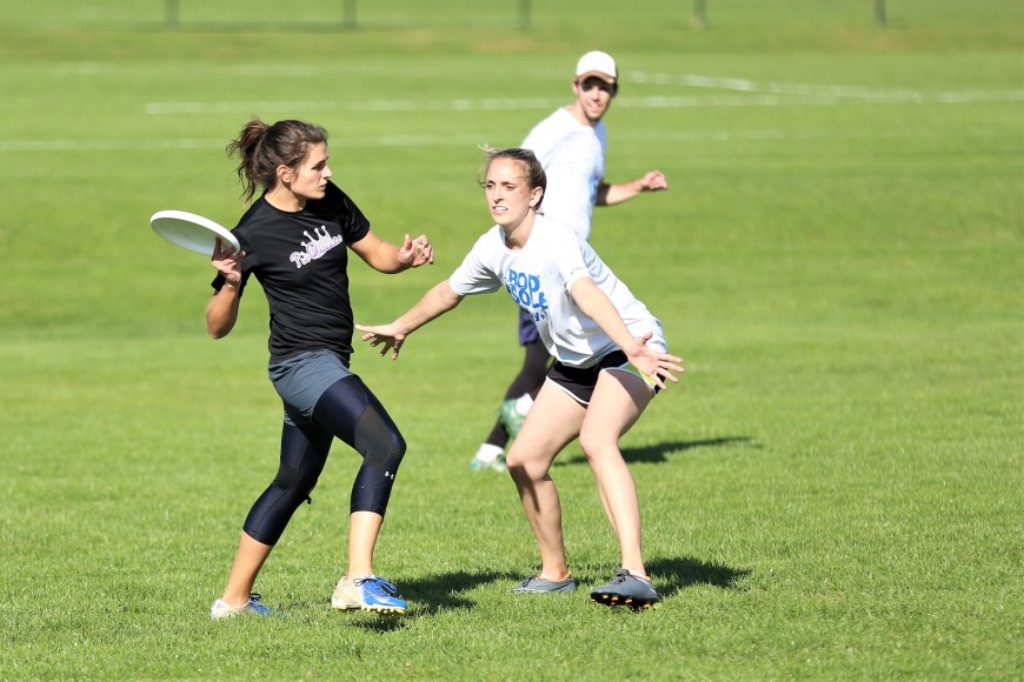 Three people are standing on a green grassy field. One white woman with brown hair in a ponytail is ready to throw a frisbee she is wearing a black t-shirt, shorts, and leggings. Standing in front of her is a white woman in a white t-shirt while a white man runs in the background also wearing a white shirt and a white hat.