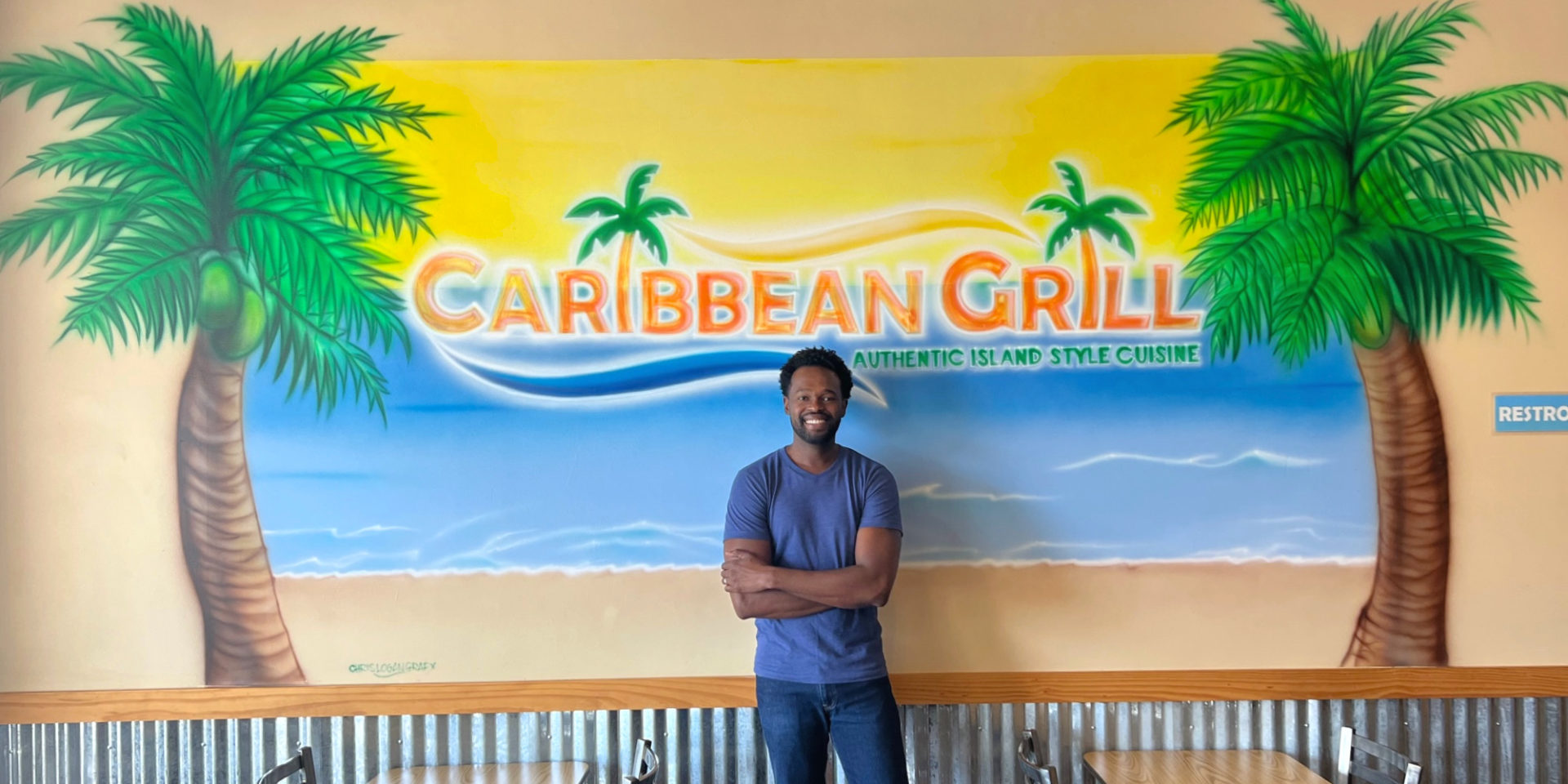 Chef Mike Harden stands inside his restaurant Caribbean Grill in Champaign, Illinois. The chef is smiling with arms folded in front of his blue t-shirt. Photo by Alyssa Buckley.