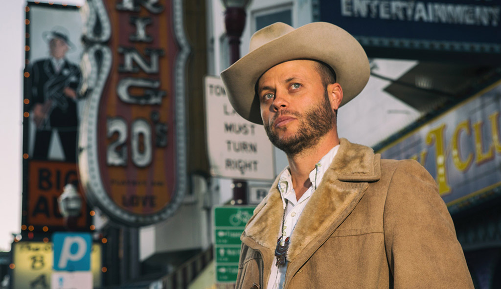 This is a photo of a person wearing a beige cowboy hat and a brown jacket with a fur collar, standing in front of a street with a lot of signs. The background consists of a street with several signs, including one that reads “BIG AL’S” and another that reads “ENTERTAINMENT”. There is also a sign that reads “PARKING” with an arrow pointing to the right. The image appears to be taken during the day.