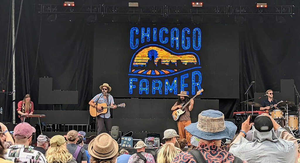 This is a photo of a band called “Chicago Farmer” performing on a stage at a music festival. The band’s name is displayed on a large screen behind the stage, written in blue and yellow on a black background. There are four band members visible on the stage, one playing the guitar, one playing the drums, one playing the keyboard, and one playing the bass. The band members are dressed in casual clothes and hats. The audience is visible in the foreground, with some people wearing hats and holding drinks.