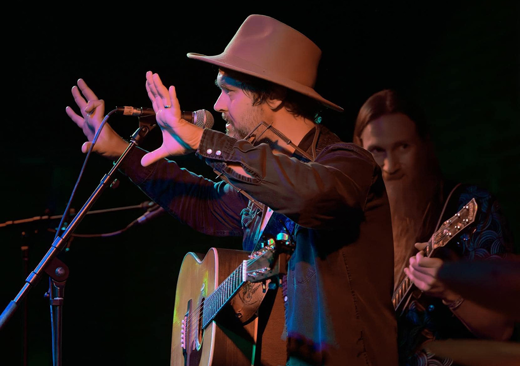 This is a photo realistic image of a musician playing guitar and singing into a microphone on a stage. The musician is wearing a cowboy hat and a black jacket, and is playing a guitar with a blue strap. The microphone is on a stand in front of the musician. The background is dark with stage lighting.